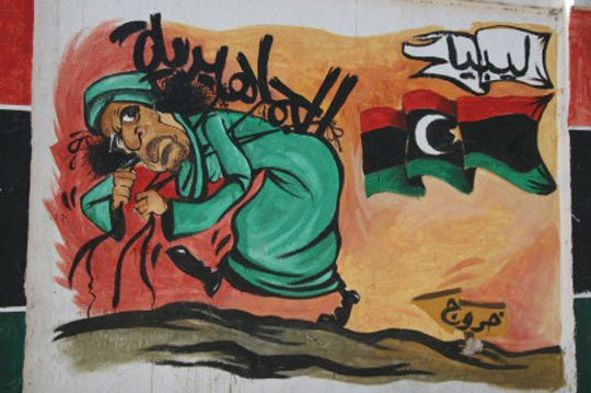 //Murals mocking Gaddafi are everywhere. This one has a sign that says “Exit”, pointing in the direction to where Gaddafi is headed. The old leader is being crushed by the weight of his Jamahiriya, the word he used to describe his so-called “state of the masses”.